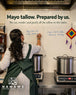 A woman in a white pinstripe shirt and green apron stands at a counter writing on a white wall next to two large metal pots on electric burners. The wall displays the NAMAWE brand logo and text about Mayo tallow balm, with handwritten details on the left and a colorful diamond-shaped hanging ornament on the right.