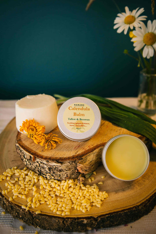 A jar of Calendula Balm with Plantago, Tallow & Beeswax on a wooden disc, alongside raw beeswax and a vase with daisy flowers in the background.