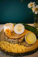 A jar of Calendula Balm with Plantago, Tallow & Beeswax on a wooden disc, alongside raw beeswax and a vase with daisy flowers in the background.